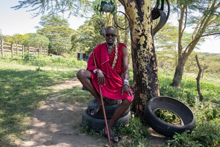 A man in a traditional Maasai shuka cloak and necklaces sits on a pile of tyres next to a tree, a stick in his hand