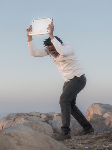 A man grimaces while holding a laptop overhead, bracing to smash it on rocks outdoors