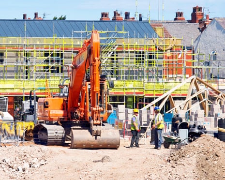 An excavator on a construction site with workers in high-visibility vests and scaffolding-clad buildings behind
