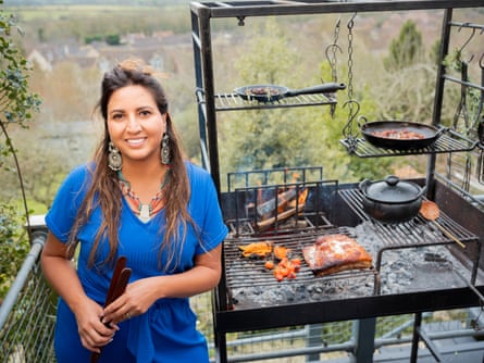 Chef Ana Ortiz is holding tongs and stands next to an outdoor fire cooking set up. There are flames at the back, and food at the front of the grill including pork and tomatoes, and recipes in pots and pans