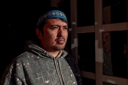 A man posing for a photograph wearing a grey patterned hoodie and a blue embroidered hat