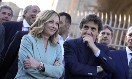 Giorgia Meloni stands among a group of formally dressed men in dark blue suits to watch the Giro d’Italia cycling race, 26 May 2024: she is wearing a light peppermint green jacket and has her blond hair loose, and is smiling as she stands with her arms folded; the light is shining on her.