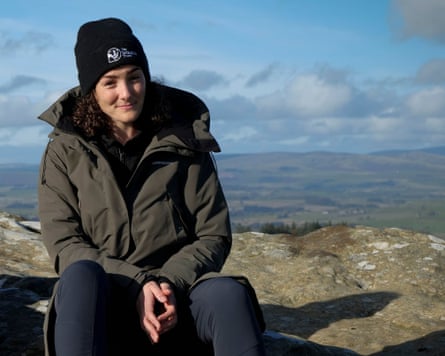 Sophie Bulgin sits on a rock on a high hillside with blue sky behind her. She has brown curly hair, and wears a black woolly hat and dark anorak.