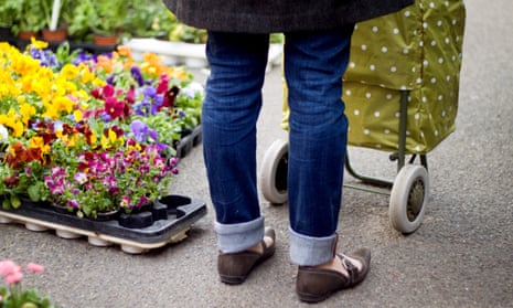A woman, seen from behind, waist down, with a spotty olive shopping trolley, standing next to some bedding plants on sale