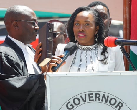Susan Kihika stands behind a podium next to a man in judicial robes holding a Bible.
