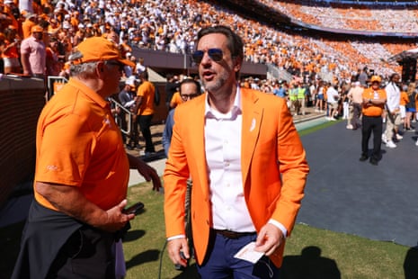 Clay Travis, in an orange blazer and sunglasses, walks across the stadium sideline talking to a man
