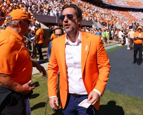 Clay Travis, in an orange blazer and sunglasses, walks across the stadium sideline talking to a man