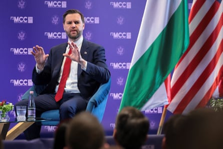 JD Vance speaks in Budapest. He is sitting in front of a purple backdrop with logos on it and the Hungarian and US flags. He is holding both hands up in front of him.
