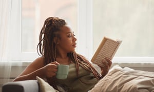 Woman reading a book<br>African young woman lying on sofa drinking coffee and reading an interesting book at home