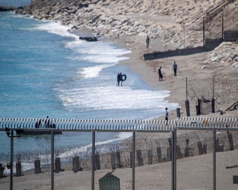 A shoreline with distant figures is seen through a high fence topped with barbed wire.