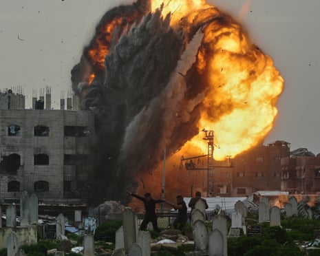 Three figures flee through a graveyard as flames and smoke surge upwards from a building in the background