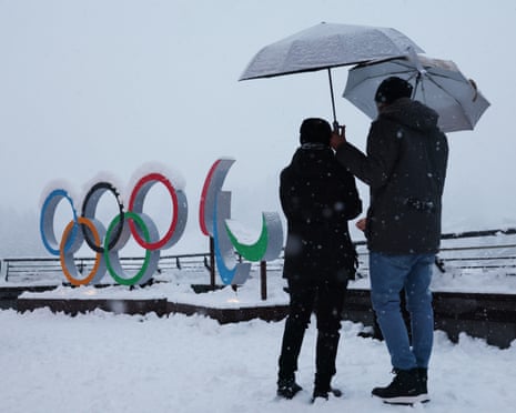Two people with umbrellas stand in the snow in front of the Olympic rings in Cortina d’Ampezzo.