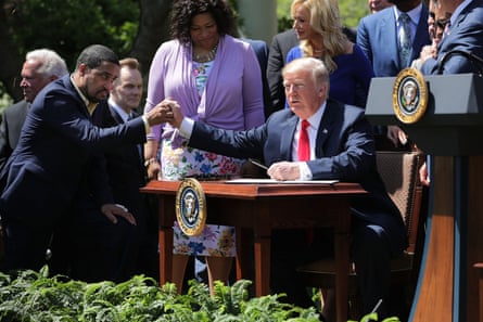 two men sitting on either side of a desk outdoors shaking hands in front of a group of people
