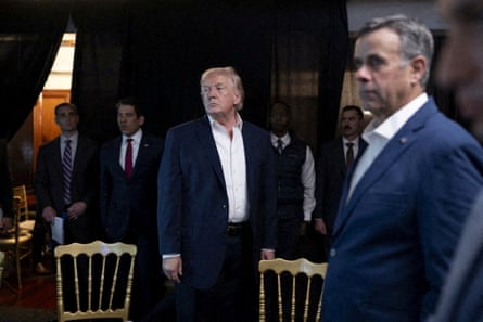 Trump in a suit but no tie stood up in a room with other men watching something on a screen