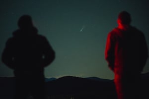 Two people with their backs turned to the camera with a white comet's tail visible in the night sky in front of them