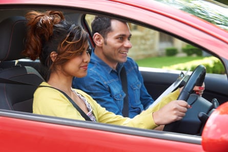 they are in a red car and seen through an open window. She wears a yellow cardigan and is gripping the steering wheel but smiling; he has a blue denim shirt and looks relaxed