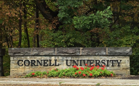 A stone wall reading "Cornell University" stands before trees, with red flowers in front, in Ithaca, N.Y