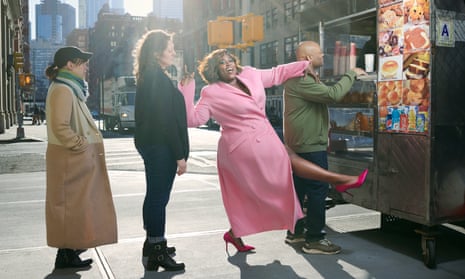Danielle Brooks standing in a burger van queue wearing a bright pink coat and red/pink satin heels; kicking out a leg, arms aloft and with a big smile