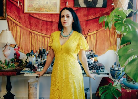 Molly Crabapple, a woman with long black hair and a yellow dress, standing against a table covered in inks and pots of pens