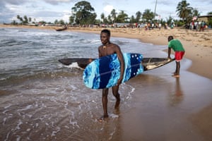 A surfer carries a surfboard while behind him a man holds a pirogue