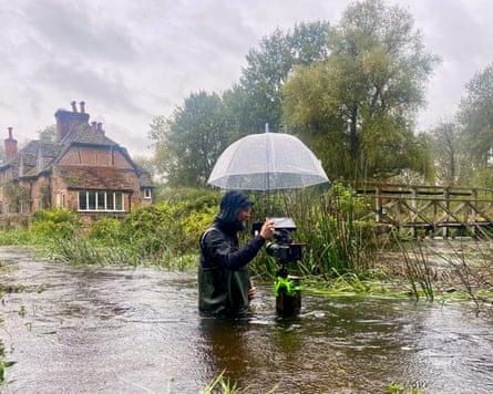 Sam Oakes, in hip-waders, stands waist-deep in a river, holding an umbrella over his camera tripod