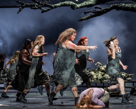 Women with long hair, streaked with dirt and wearing torn tunics, march across a stage looking angry as a woman kneels on the floor, head in her hands