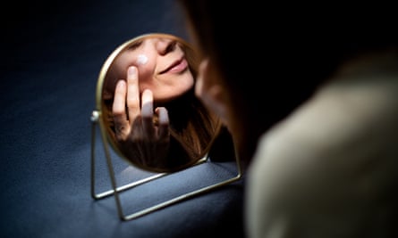 Woman looking in the mirror at herself, applying moisturizer on her face and smiling