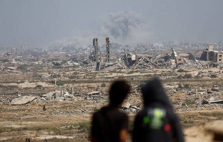 Two people look towards a landscape of rubble, from where smoke is rising in the distance