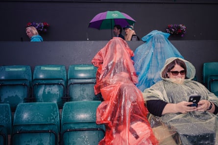 Visitors wearing plastic ponchos sit in the stands of the court while play is suspended