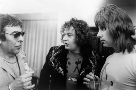 Black and white photo showing three actors dressed in character as 80s rockers, standing in a group talking