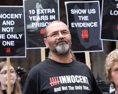 Andrew Malkinson in glasses and a black T-shirt stands at a protest holding signs behind him