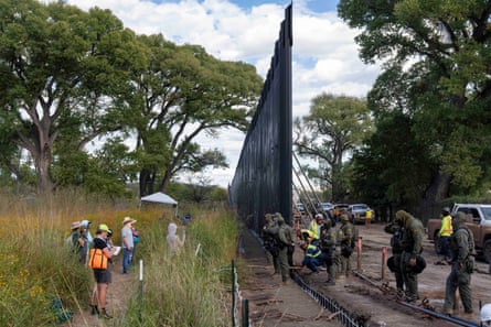A line of people face a line of officers standing by border wall outside