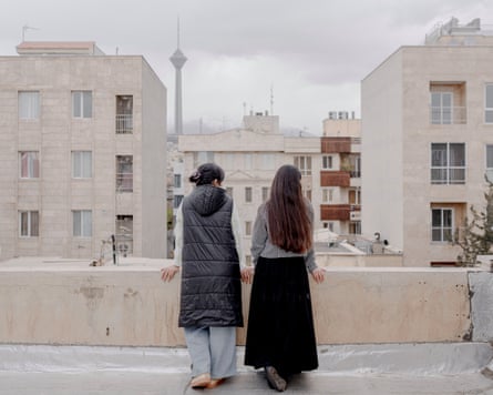 Two women looking out from a balcony or low rooftop wall towards similar light-coloured buildings
