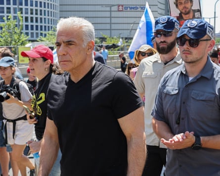 Yair Lapid walks in a crowd at a rally in Tel Aviv; he is in his early 60s with white slicked-black hair and wears a black T-shirt.