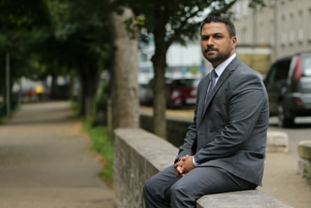 Andy George sits on a low stone wall at the edge of a park with tall stone buildings in the background; he is out of uniform and wears a grey suit and tie.