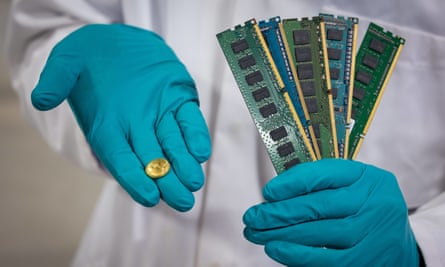 a person in a white lab coat holds a small gold piece in one hand and a bunch of waste circuit boards in the other. Both their hands are clad in blue surgical-style rubber gloves