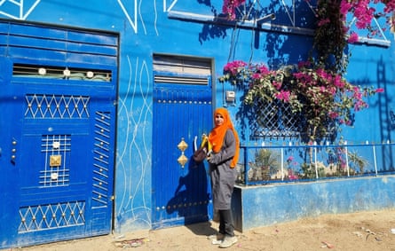 A young woman in a dustcoat and an orange hijab stands at the front door of a blue house with bougainvillaea outside