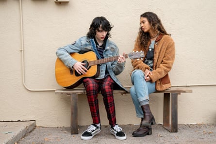 Wolfhard wearing a denim jacket playing the guitar, sitting next to Boe, who is wearing a tan jacket