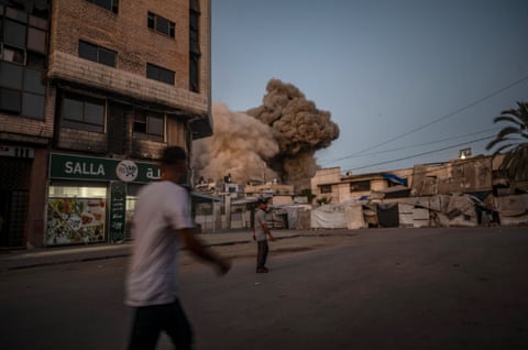 People on a street in Gaza as a large plume of brown smoke rises from buildings.