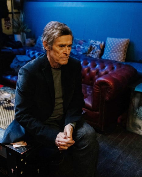Actor Willem Dafoe wearing a dark blue jacket and grey top sits on the edge of a coffee table, hands folded