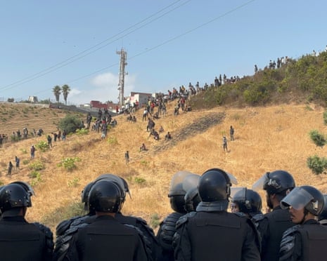 In the foreground, a group of men in black uniforms and helmets observe a group of men on a parched grassy hillside in the distance.
