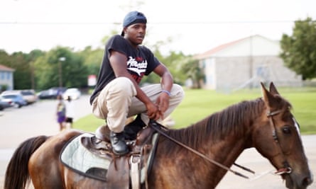 A young man sitting in a squat position on the saddle of a horse.