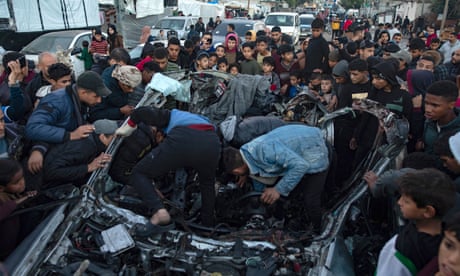 A crowd of people surround a car with its roof blown off on a street, with some people clambering into the wreckage.