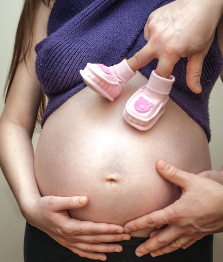 A pregnant woman holding her exposed belly. A hand with two baby shoes on its fingers is walking on the belly.