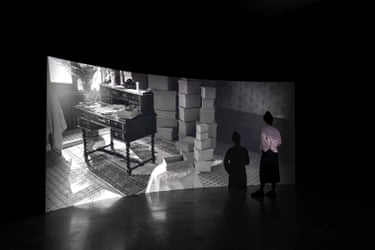 In Hoda Afshar’s “The Fold”, figures view a projected black-and-white room with a desk and stacked boxes