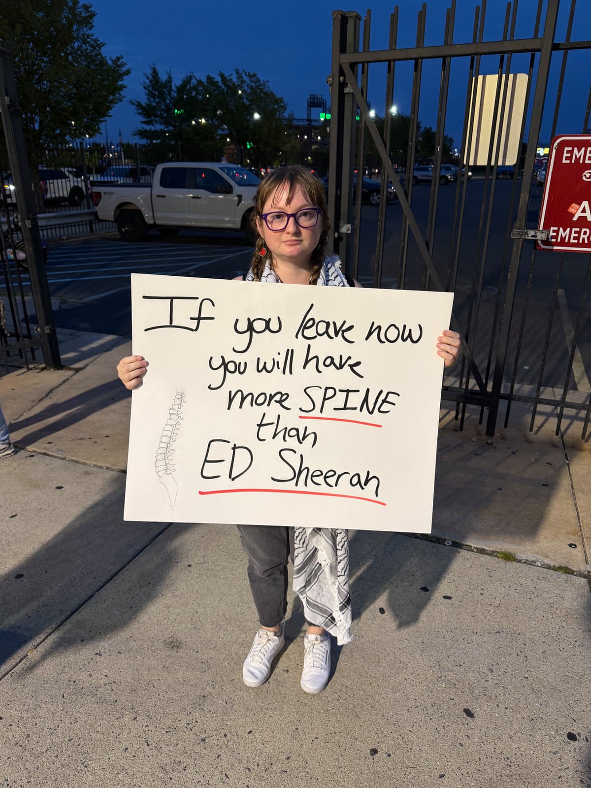 a woman holds a sign outside a stadium at nighttime reading ‘if you leave now you will have more spine than Ed Sheeran’