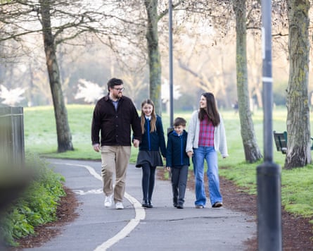A family, dad, daughter, son and mum walk in a line holding hands through a park.