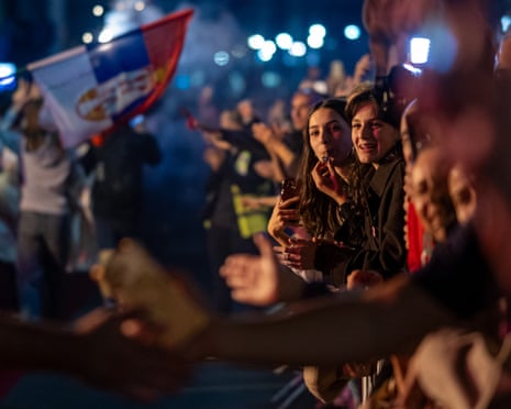 Student protesters smile at a crowded night rally in Belgrade, one holding a phone