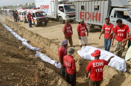 People in red T-shirts stand around a body bag near a line of body bags in a graveyard, burying the dead.
