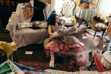 Wearing a frilly dress and big heels, she lies on the floor of a messy living room reading a newspaper